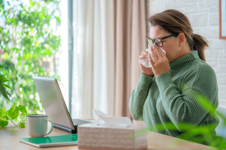 Woman sneezing into a tissue while sitting at a desk with a laptop at home.